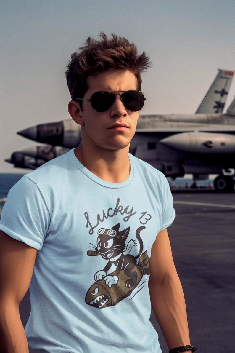 May include: A light blue t-shirt featuring a graphic of a black cat wearing goggles and smoking a cigar while riding a brown bomb. The text Lucky 13 is printed above the cat in a cursive font. The shirt has short sleeves and a crew neckline.