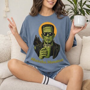 Może przedstawiać: Niebieski t-shirt z grafiką potwora Frankensteina w okularach przeciwsłonecznych, pijącego zielony nap&oacute;j z plastikowego kubka ze słomką. Za postacią znajduje się ż&oacute;łte koło i sylwetka zamku. Poniżej widnieje napis Matcha Monster.