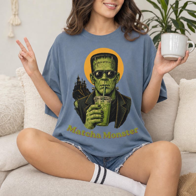 Może przedstawiać: Niebieski t-shirt z grafiką potwora Frankensteina w okularach przeciwsłonecznych, pijącego zielony nap&oacute;j z plastikowego kubka ze słomką. Za postacią znajduje się ż&oacute;łte koło i sylwetka zamku. Poniżej widnieje napis Matcha Monster.
