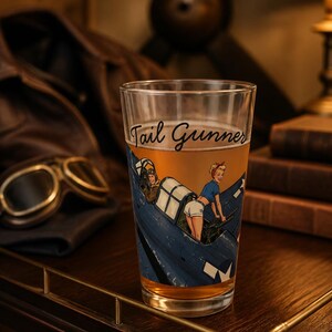 May include: A clear glass pint glass filled with a golden liquid, featuring a printed graphic of a blue military aircraft with a pilot and a person in a blue shirt and white shorts operating a machine gun, with the text Tail Gunner in script.