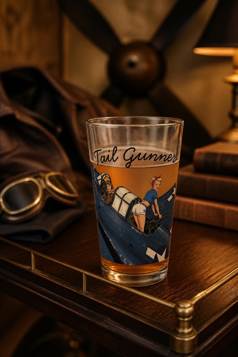 May include: A clear glass pint glass filled with a golden liquid, featuring a printed graphic of a blue military aircraft with a pilot and a person in a blue shirt and white shorts operating a machine gun, with the text Tail Gunner in script.