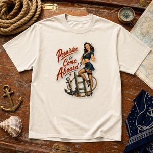 Op de afbeelding: Een cr&egrave;mekleurig t-shirt met korte mouwen en een afbeelding van een zeeman in een marineblauw uniform bij een ladder en een anker. De tekst luidt Permission to Come Aboard? in rode letters. Het ontwerp bevat een touwaccent.