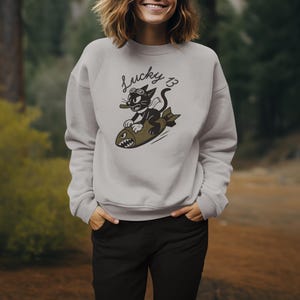 May include: A light gray sweatshirt featuring a cartoon cat riding a bomb, with the text "Lucky 13" above. The cat wears goggles and holds a cigar. The sweatshirt is paired with black pants.