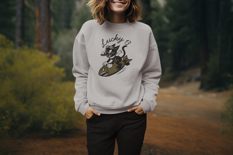 May include: A light gray sweatshirt featuring a cartoon cat riding a bomb, with the text "Lucky 13" above. The cat wears goggles and holds a cigar. The sweatshirt is paired with black pants.