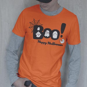 May include: Orange t-shirt with the word "Boo!" in a black bubble font, featuring ghost illustrations and a spiderweb. The text "Happy Halloween" is below. The shirt is worn over a gray long-sleeved shirt.