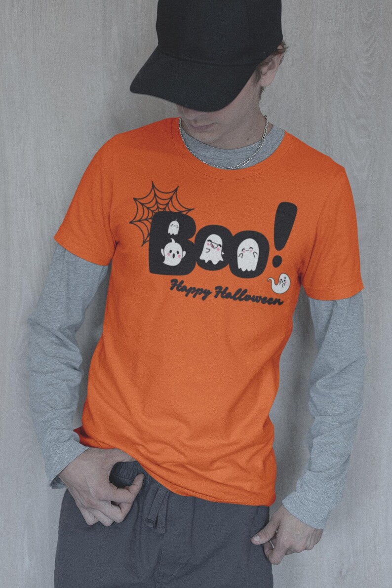May include: Orange t-shirt with the word "Boo!" in a black bubble font, featuring ghost illustrations and a spiderweb. The text "Happy Halloween" is below. The shirt is worn over a gray long-sleeved shirt.