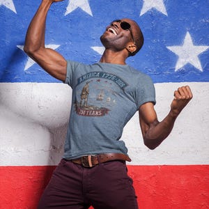 May include: A heather blue t-shirt featuring a graphic print of a historical figure, sailing ships, and the text America 1776-2026 250 Years. The design includes a banner and stars, centred on the chest of the short-sleeved crew neck garment.