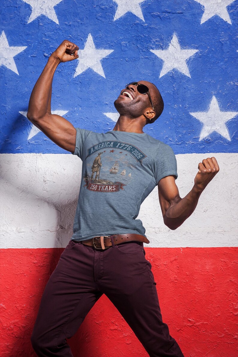 May include: A heather blue t-shirt featuring a graphic print of a historical figure, sailing ships, and the text America 1776-2026 250 Years. The design includes a banner and stars, centred on the chest of the short-sleeved crew neck garment.