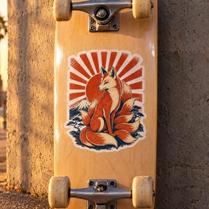 May include: A light-colored wooden skateboard deck featuring a central graphic of a multi-tailed fox sitting before a large red sun, stylized mountain peaks, and pine trees. The skateboard includes silver metal trucks and off-white wheels.