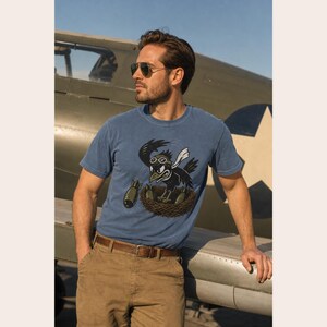 Puede incluir: Una camiseta azul de manga corta con un estampado gr&aacute;fico de un p&aacute;jaro negro con gafas de aviador y una bufanda blanca, sentado en un nido con tres bombas de color verde oliva. El p&aacute;jaro tiene una expresi&oacute;n decidida y suelta una bomba.