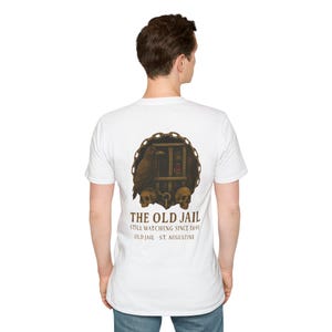 May include: White t-shirt featuring a graphic of a jail cell with a raven, skulls, and the text "THE OLD JAIL STILL WATCHING SINCE 1891 OLD JAIL ST. AUGUSTINE."