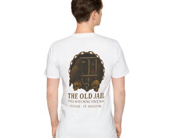 May include: White t-shirt featuring a graphic of a jail cell with a raven, skulls, and the text "THE OLD JAIL STILL WATCHING SINCE 1891 OLD JAIL ST. AUGUSTINE."
