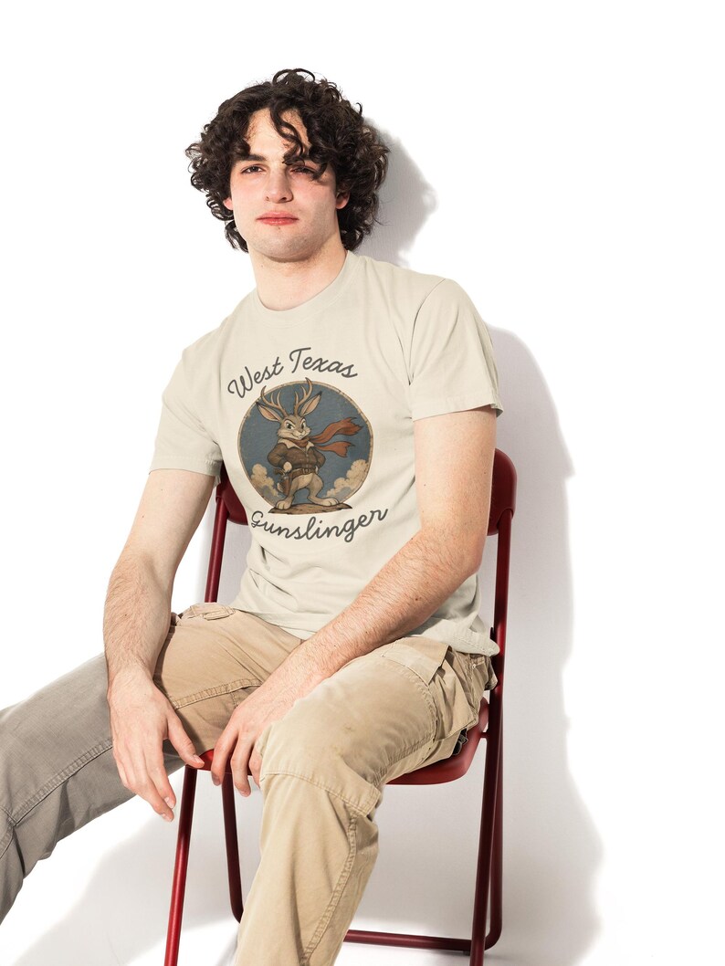 May include: A cream-colored short-sleeve t-shirt featuring a circular graphic of a jackalope wearing a brown vest and a red scarf. The text "West Texas Gunslinger" is printed in a dark script font around the circular illustration.