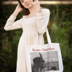 May include: Cream-colored tote bag with the text "Stone Guardian of the City of Love" in red. The bag features a black and white image of a statue and a cityscape. The bag is held by a person wearing a cream-colored dress.