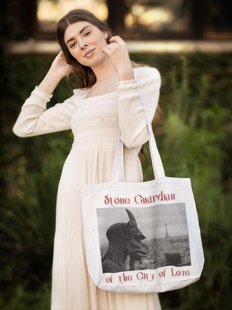 May include: Cream-colored tote bag with the text "Stone Guardian of the City of Love" in red. The bag features a black and white image of a statue and a cityscape. The bag is held by a person wearing a cream-colored dress.