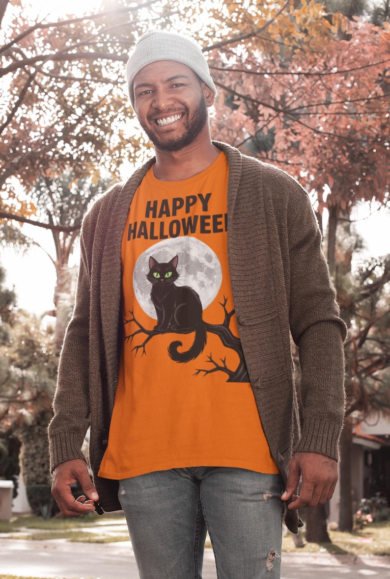 Może przedstawiać: Pomarańczowa koszulka z czarnym kotem na gałęzi przed pełnią księżyca. Tekst "HAPPY HALLOWEEN!" jest czarny. Osoba ma na sobie szarą czapkę, brązowy kardigan i niebieskie dżinsy. Motyw Halloween.