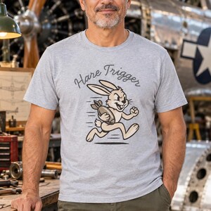 Puede incluir: Una camiseta gris jaspeado de manga corta con un gr&aacute;fico de un conejo de dibujos animados corriendo mientras sostiene una bomba. Sobre el conejo, el texto "Hare Trigger" aparece en una fuente cursiva oscura con contornos negros.