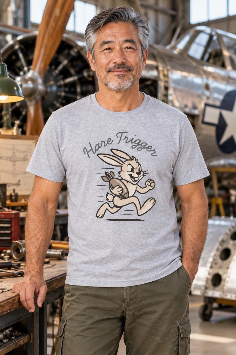 Puede incluir: Una camiseta gris jaspeado de manga corta con un gr&aacute;fico de un conejo de dibujos animados corriendo mientras sostiene una bomba. Sobre el conejo, el texto "Hare Trigger" aparece en una fuente cursiva oscura con contornos negros.