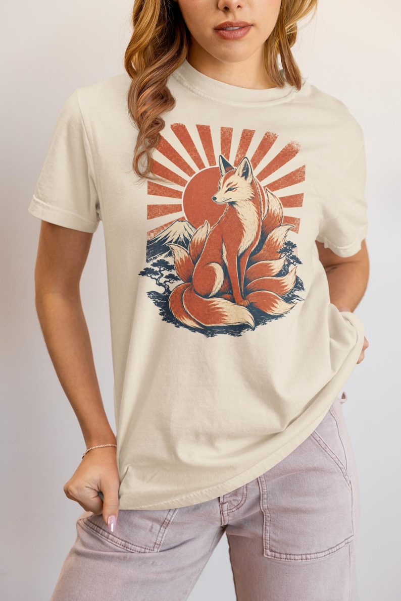 May include: A cream-colored short-sleeve t-shirt featuring a graphic print of a multi-tailed fox sitting in front of a large red sun, a mountain peak, and stylized trees. The design uses a distressed, vintage-inspired color palette of red and blue.