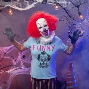 May include: A person in a clown costume with a red wig, white face, and a shirt that says "PRETTY FUNNY". The clown is wearing black gloves and is surrounded by cobwebs and Halloween decorations.