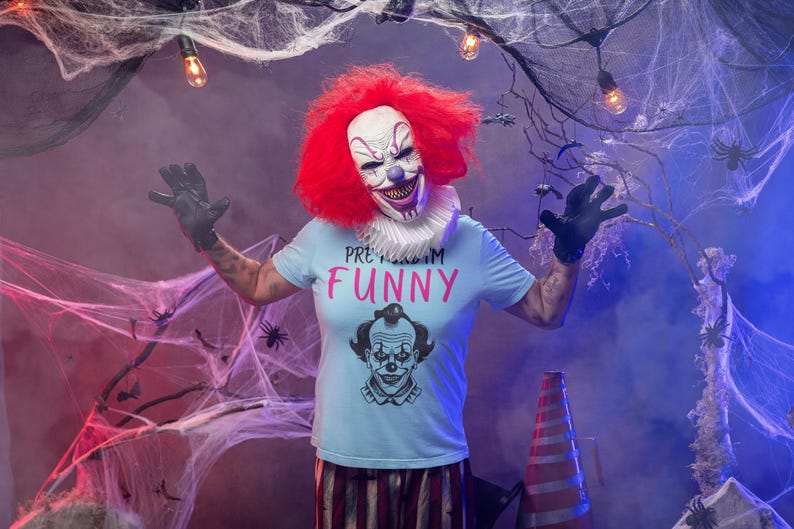 May include: A person in a clown costume with a red wig, white face, and a shirt that says "PRETTY FUNNY". The clown is wearing black gloves and is surrounded by cobwebs and Halloween decorations.