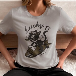 May include: A light gray short-sleeved t-shirt featuring a black and olive green graphic of a cartoon cat wearing goggles and a scarf while riding a bomb. The text Lucky 13 is printed in black above the cat, which is holding a cigar in its mouth.