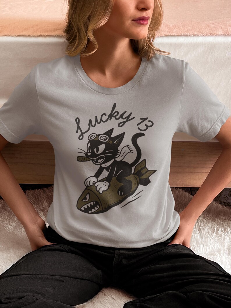 May include: A light gray short-sleeved t-shirt featuring a black and olive green graphic of a cartoon cat wearing goggles and a scarf while riding a bomb. The text Lucky 13 is printed in black above the cat, which is holding a cigar in its mouth.