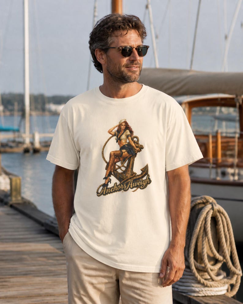 May include: A cream-colored short-sleeve t-shirt featuring a graphic print of a person in a sailor outfit sitting on a large metal anchor with a rope. The text "Anchors Aweigh" is printed in a dark script font below the anchor illustration.