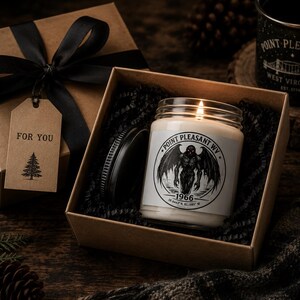May include: A lit candle in a glass jar with a black lid, featuring a label with a winged creature illustration, the text Point Pleasant WV, 1966, and coordinates. It is nestled in a brown gift box with black filler, a black ribbon, and a tag reading FOR YOU.