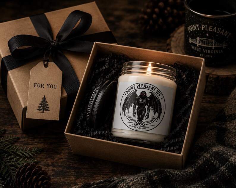 May include: A lit candle in a glass jar with a black lid, featuring a label with a winged creature illustration, the text Point Pleasant WV, 1966, and coordinates. It is nestled in a brown gift box with black filler, a black ribbon, and a tag reading FOR YOU.
