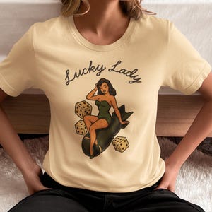 May include: A cream-colored short-sleeve t-shirt featuring a graphic print of a pin-up style figure sitting on a dark green bomb, surrounded by three dice. The text "Lucky Lady" is printed in a black cursive script above the central illustration.