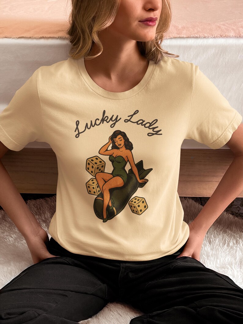 May include: A cream-colored short-sleeve t-shirt featuring a graphic print of a pin-up style figure sitting on a dark green bomb, surrounded by three dice. The text "Lucky Lady" is printed in a black cursive script above the central illustration.