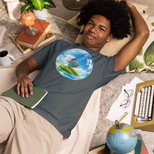 May include: A dark gray short-sleeve t-shirt featuring a circular graphic print on the chest. The graphic depicts a blue discus fish swimming inside a transparent bubble containing a small green grassy island under a bright blue sky with clouds.