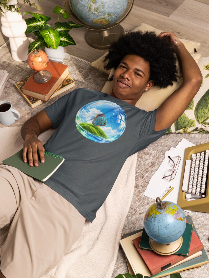 May include: A dark gray short-sleeve t-shirt featuring a circular graphic print on the chest. The graphic depicts a blue discus fish swimming inside a transparent bubble containing a small green grassy island under a bright blue sky with clouds.