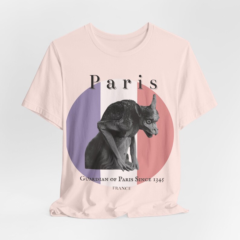 May include: A light pink t-shirt featuring a black and white gargoyle illustration. The design includes a circular graphic with the colors of the French flag, the word "Paris", and the text "Guardian of Paris Since 1345 FRANCE".