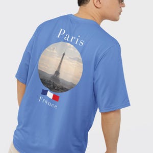 May include: A blue t-shirt featuring a circular graphic of the Eiffel Tower in Paris, France. The word "Paris" is printed above the graphic, and "France" with the French flag is below. The shirt is a solid blue color.