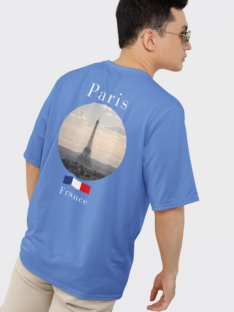 May include: A blue t-shirt featuring a circular graphic of the Eiffel Tower in Paris, France. The word "Paris" is printed above the graphic, and "France" with the French flag is below. The shirt is a solid blue color.