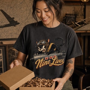 以下が含まれることがあります： チャコールグレーの半袖Tシャツ。飛行帽とゴーグルを身につけ、爆弾にまたがった黒猫のグラフィックが描かれています。猫の下には黄色の筆記体で「Nine Lives」という文字と、3つの小さな赤と白の太陽のアイコンがプリントされています。