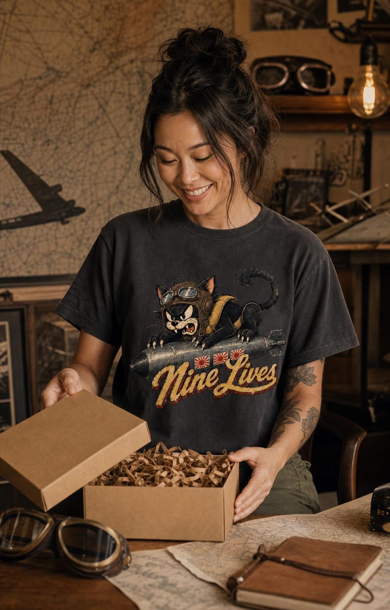 以下が含まれることがあります： チャコールグレーの半袖Tシャツ。飛行帽とゴーグルを身につけ、爆弾にまたがった黒猫のグラフィックが描かれています。猫の下には黄色の筆記体で「Nine Lives」という文字と、3つの小さな赤と白の太陽のアイコンがプリントされています。