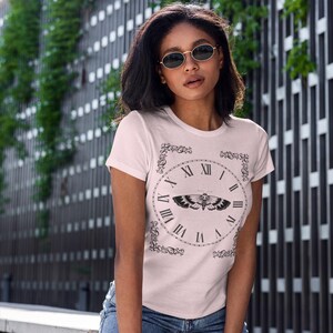 May include: Pale pink t-shirt featuring a black clock design with Roman numerals and a moth illustration. The shirt is a short-sleeved crew neck style. The model is wearing blue jeans and sunglasses.