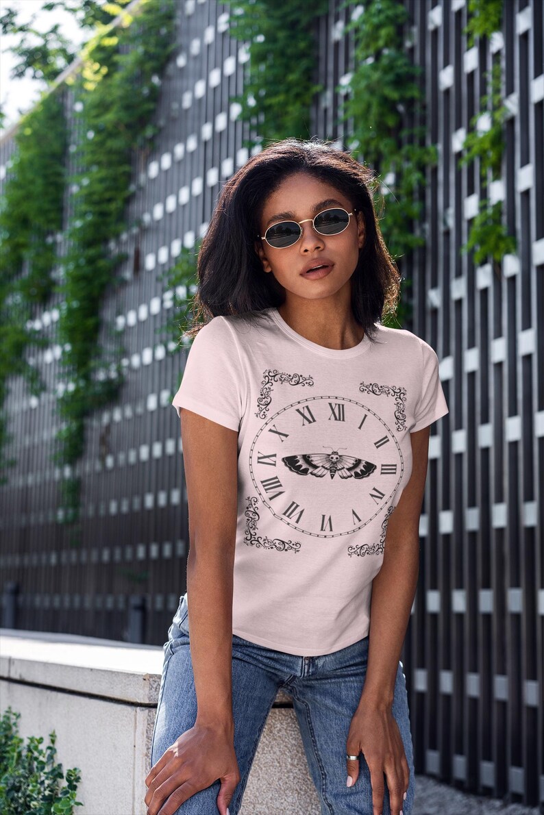 May include: Pale pink t-shirt featuring a black clock design with Roman numerals and a moth illustration. The shirt is a short-sleeved crew neck style. The model is wearing blue jeans and sunglasses.