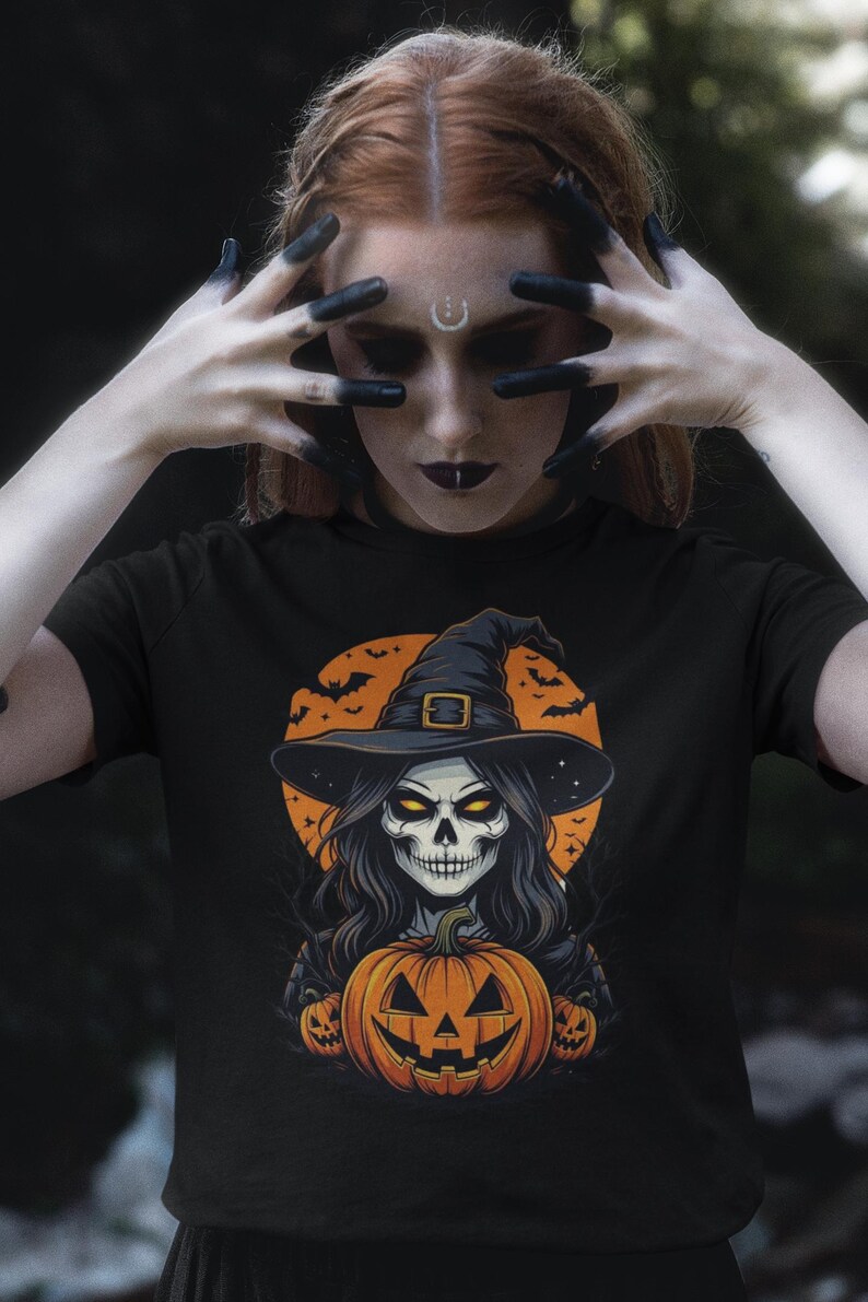 May include: Black t-shirt featuring a Halloween-themed graphic. The design includes a skull wearing a witch hat, long hair, and a carved pumpkin. The background has bats and a full moon. The person in the image has black painted fingers.