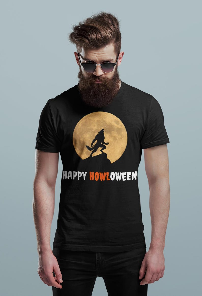 May include: Black t-shirt with a silhouette of a howling wolf in front of a full moon. The text "HAPPY HOWLOWEEN" is printed in white and orange. A Halloween-themed design, perfect for the spooky season.