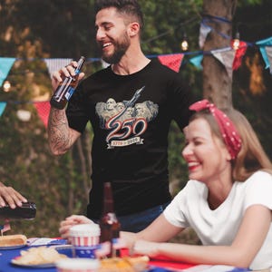 May include: A black short-sleeved t-shirt featuring a graphic of Mount Rushmore with a jet flying overhead, the number 250 in red, white, and blue, and the text 1776-2026 on a banner. The design is centred on the chest of the garment.