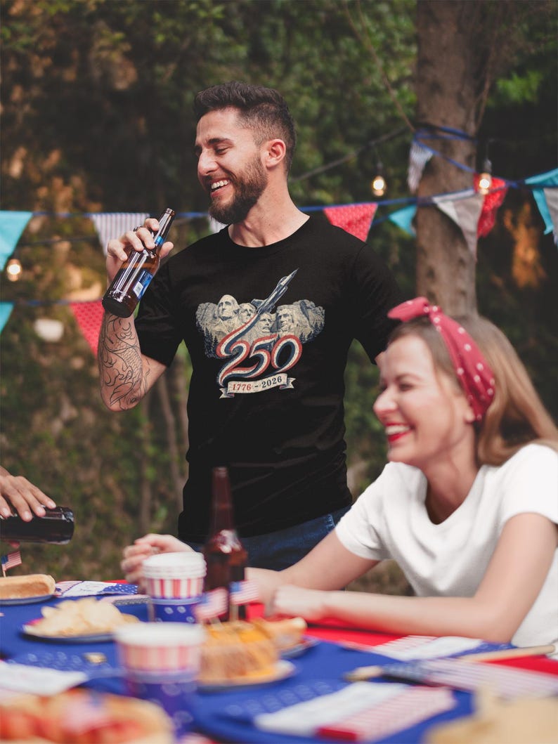 May include: A black short-sleeved t-shirt featuring a graphic of Mount Rushmore with a jet flying overhead, the number 250 in red, white, and blue, and the text 1776-2026 on a banner. The design is centred on the chest of the garment.