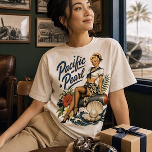 May include: A white short-sleeve t-shirt featuring a graphic print of a blonde woman in a sailor outfit sitting on a torpedo, surrounded by tropical flowers, a large open oyster shell with a pearl, and the text Pacific Pearl in blue script.