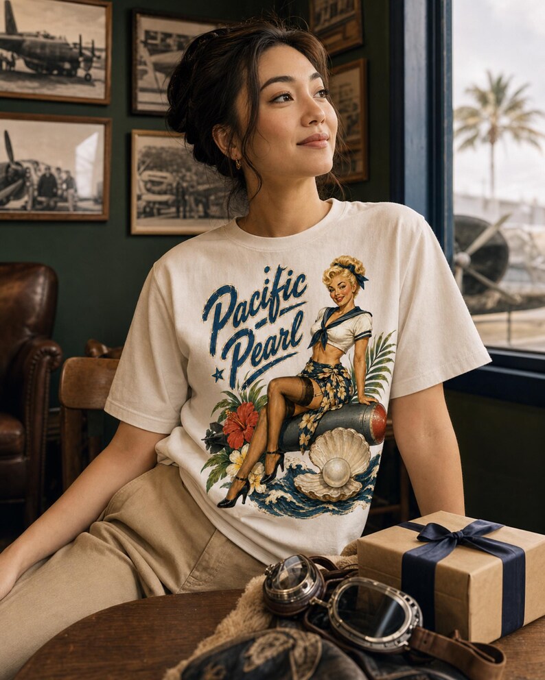 May include: A white short-sleeve t-shirt featuring a graphic print of a blonde woman in a sailor outfit sitting on a torpedo, surrounded by tropical flowers, a large open oyster shell with a pearl, and the text Pacific Pearl in blue script.