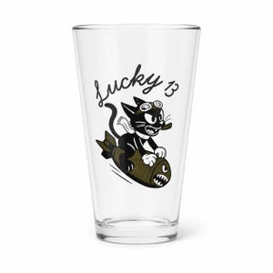 May include: A clear glass pint tumbler featuring a black and olive green graphic of a cartoon cat wearing goggles and a scarf while riding a bomb. The text Lucky 13 is printed in black script above the cat. The design has a vintage aesthetic.