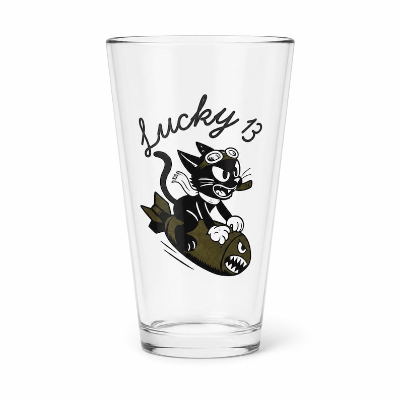 May include: A clear glass pint tumbler featuring a black and olive green graphic of a cartoon cat wearing goggles and a scarf while riding a bomb. The text Lucky 13 is printed in black script above the cat. The design has a vintage aesthetic.