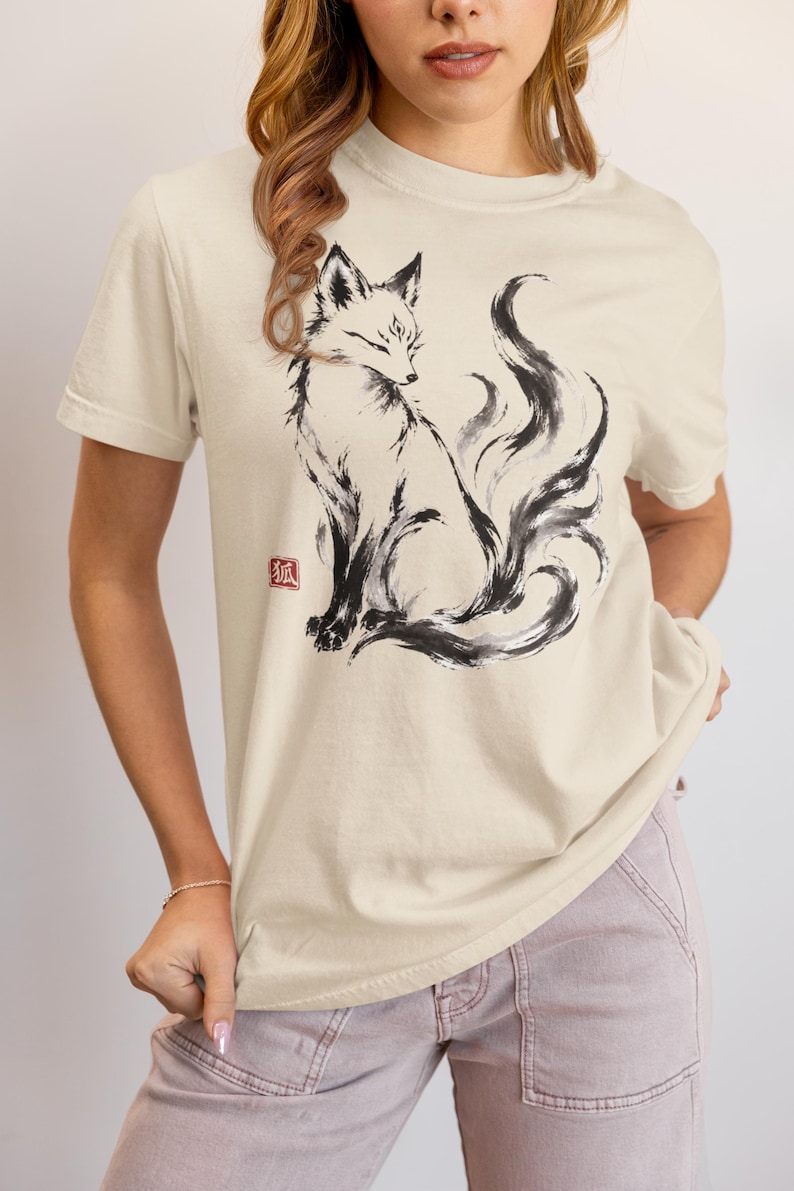 K&ouml;nnte beinhalten: Ein beiges Kurzarm-T-Shirt mit einer schwarzen Tuschezeichnung eines Fuchses mit mehreren Schw&auml;nzen. Das Motiv enth&auml;lt einen kleinen roten quadratischen Stempel mit dem japanischen Kanji-Zeichen f&uuml;r Fuchs, 狐, links neben den Vorderpfoten des Fuchses.